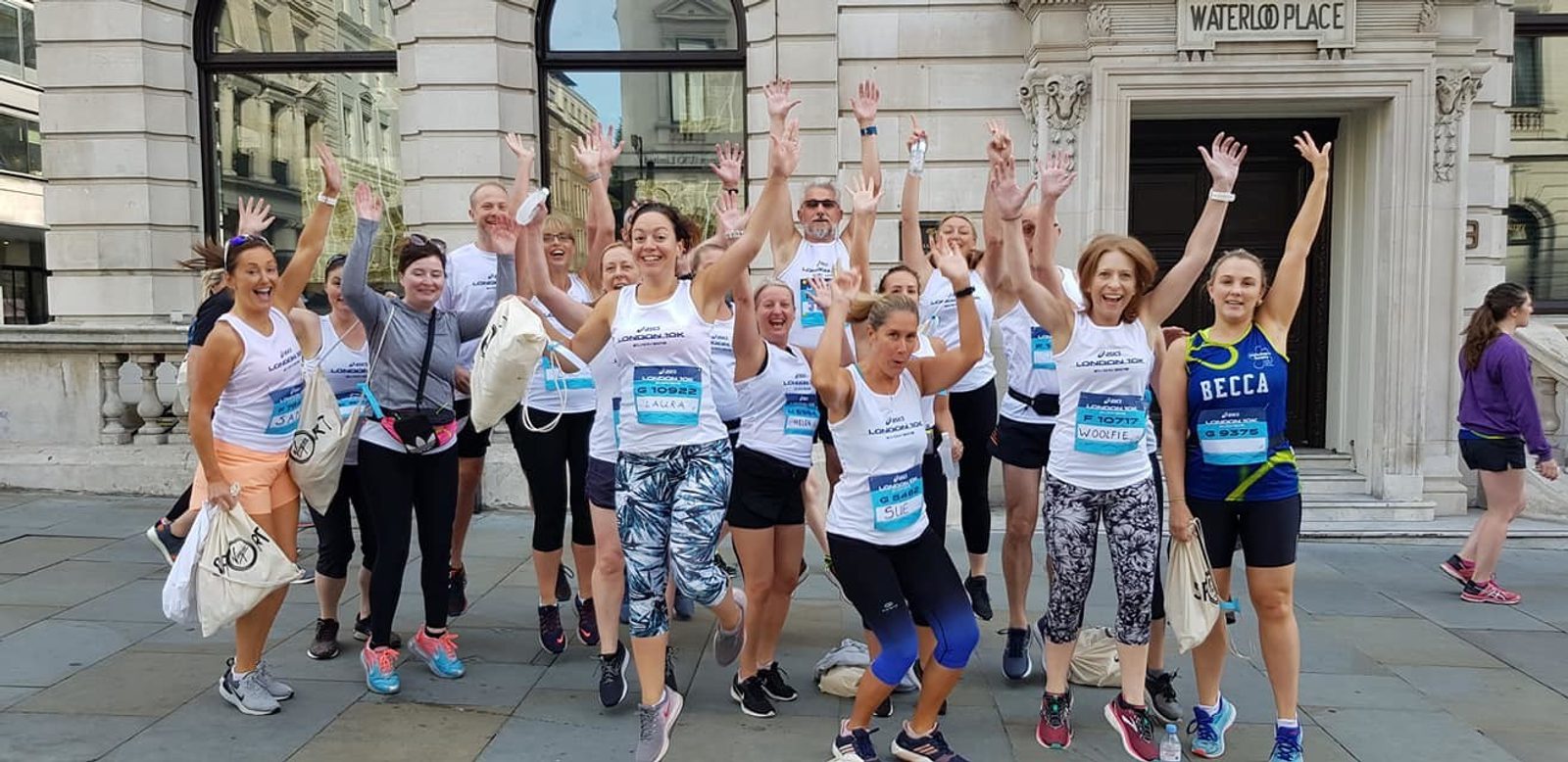A group of people in athletic wear are joyfully posing and jumping together in front of a historic building with "Waterloo Place" inscribed above the entrance. Some are holding bags, and many are wearing race bibs and shirts, suggesting they have participated in an event.