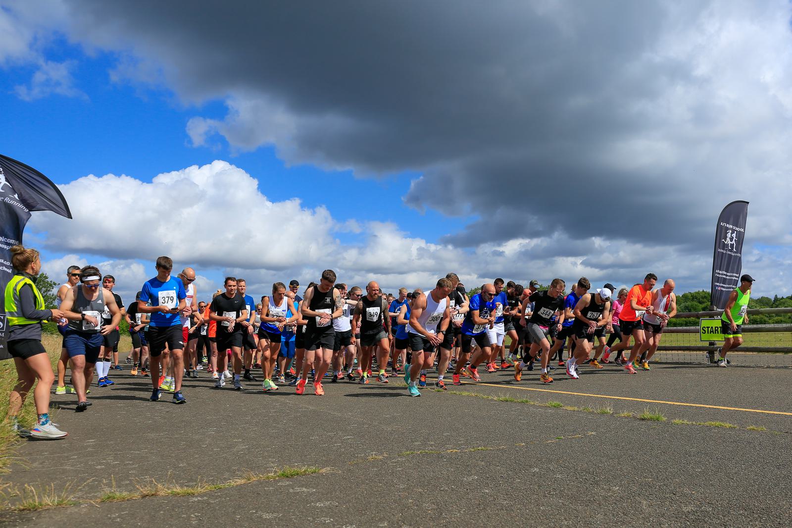 A large group of runners at the starting line of a race on a paved road under a blue sky with clouds. An official in a neon yellow vest is on the left, and large banners mark the start. Runners are wearing various athletic clothing and bib numbers.