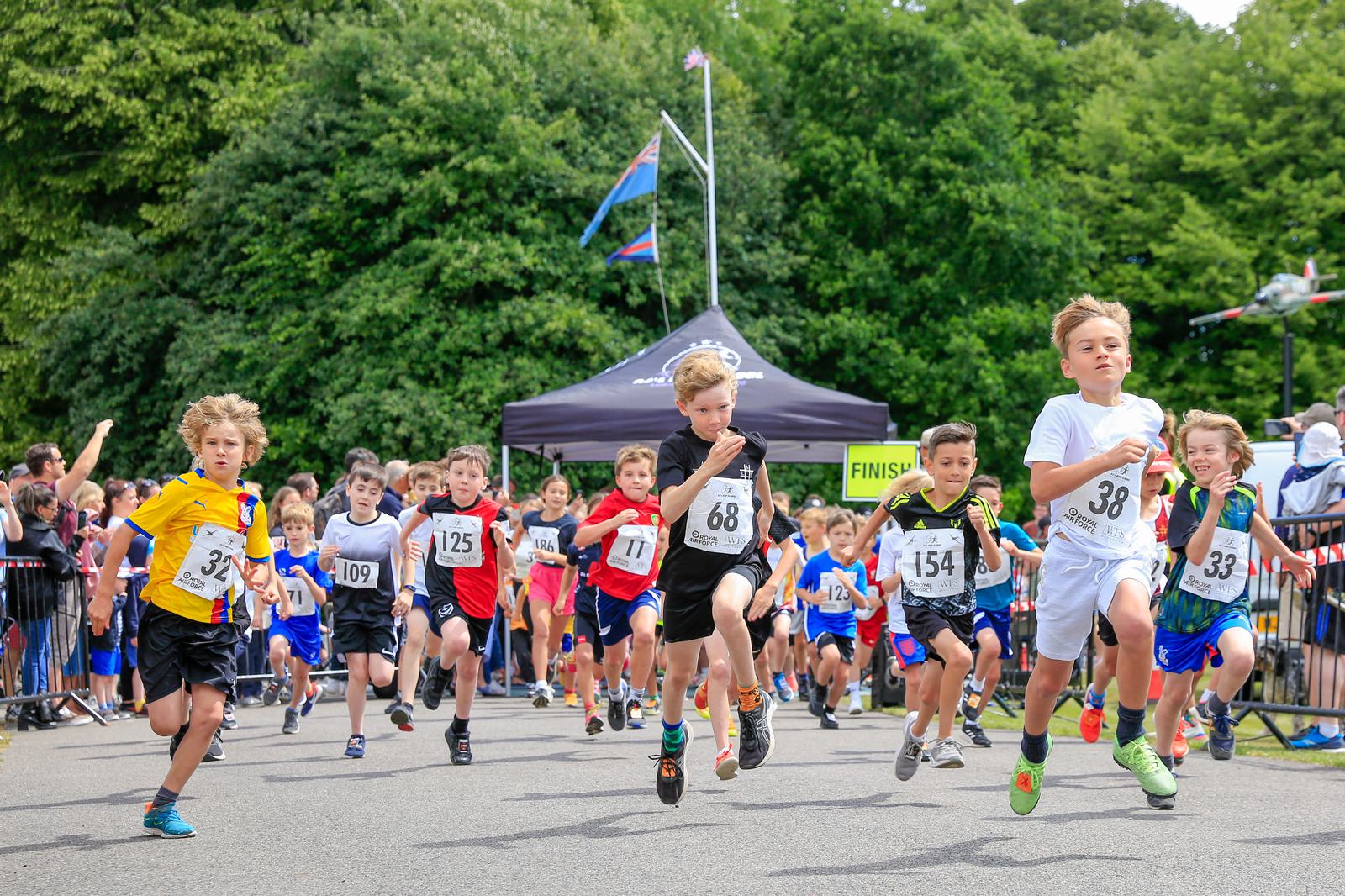 A group of children wearing numbered bibs race towards the finish line in an outdoor event. They run on a paved path lined with spectators behind barriers. Green trees are visible in the background along with a black tent and flags.