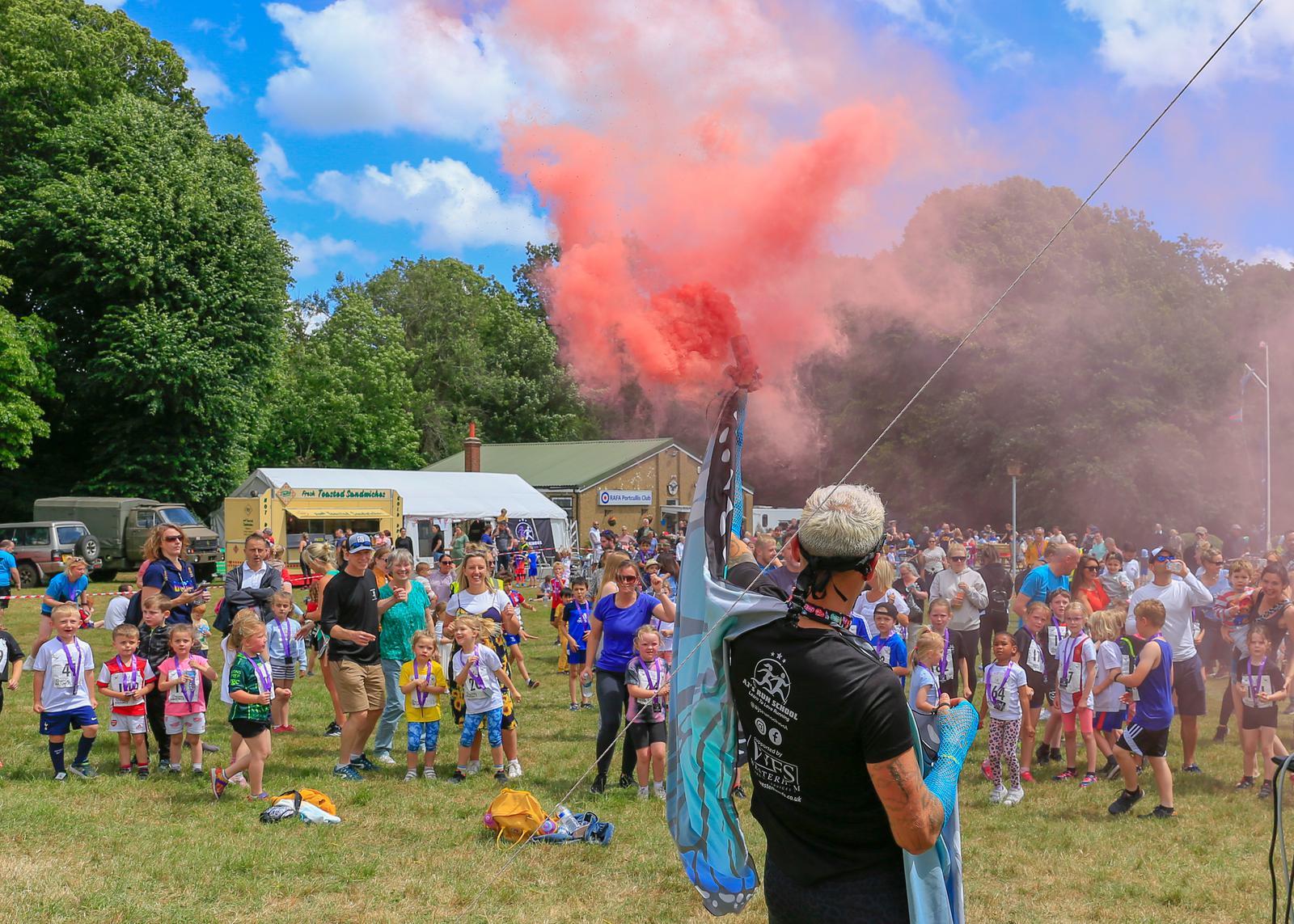 A vibrant outdoor event with children and adults gathered in a grassy area. A person in the foreground releases colorful smoke into the air while holding a flag. Tents and trees are visible in the background under a blue sky with clouds.