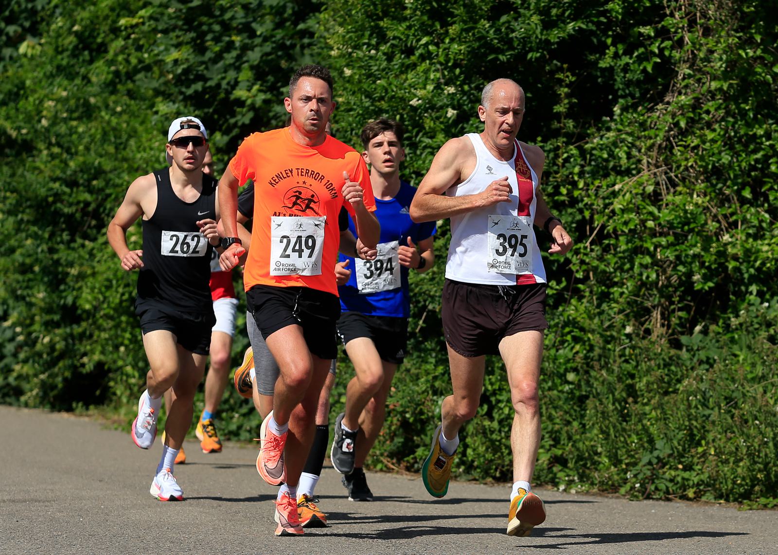 Four runners are competing in a race, running on a paved path lined with greenery. Each has a race number pinned to their shirts. The lead runner wears a white top and the next runner wears an orange top. It appears to be a sunny day.