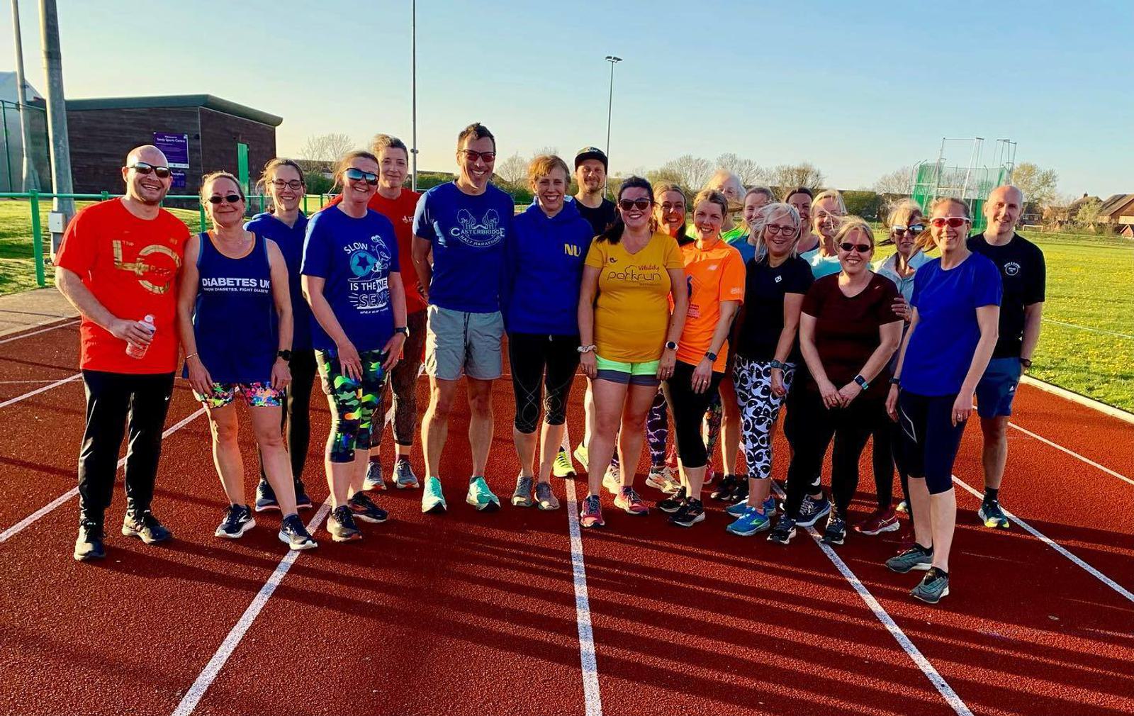 A group of people are posing on an outdoor running track on a sunny day. They are dressed in athletic wear, with some wearing bright colors. Everyone is smiling and standing close together in a friendly manner. Trees and sports equipment are visible in the background.