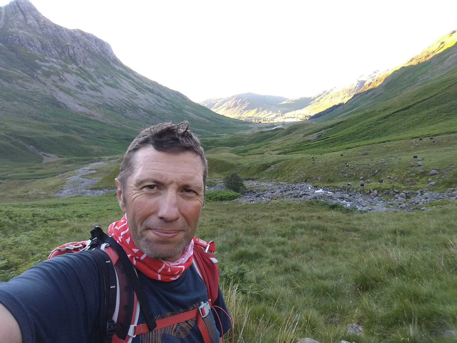 A man wearing a red and white neck gaiter and a backpack is taking a selfie in a lush, green valley with rolling hills and a distant village. The sky is clear, and sunlight illuminates the landscape.