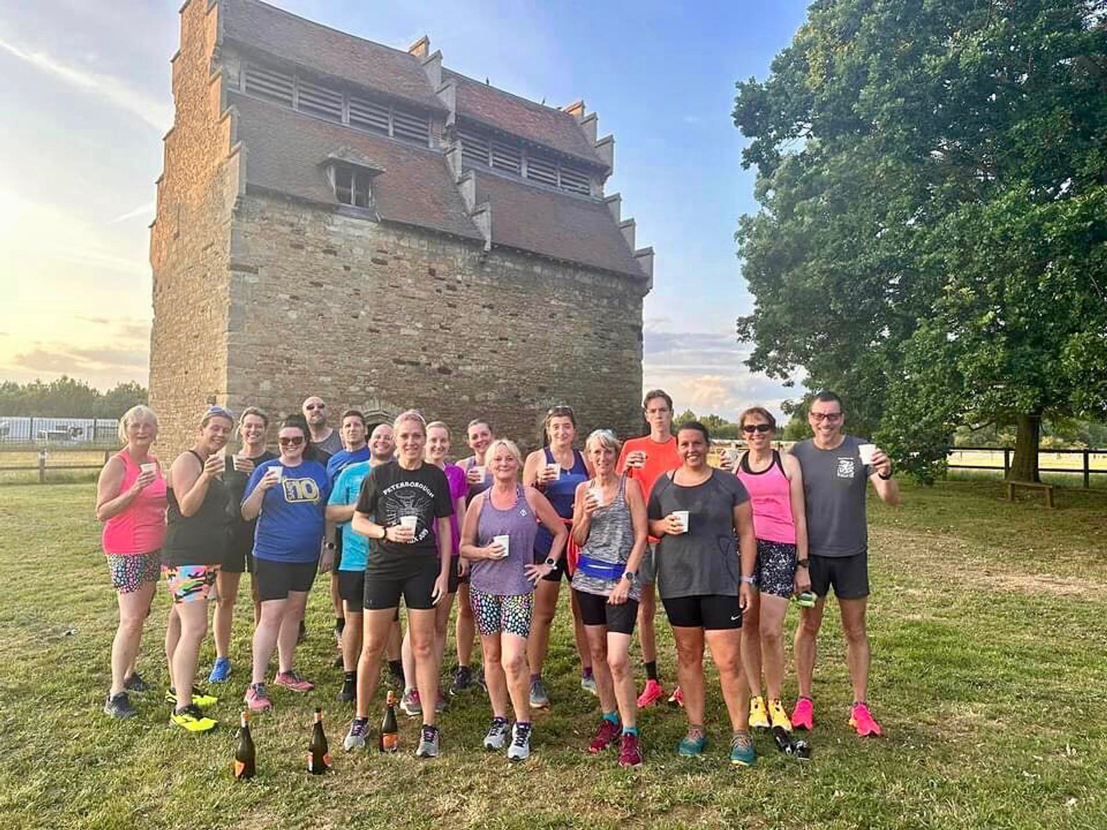 A group of people in athletic wear pose outdoors, holding drinks and smiling at the camera. They stand on grassy ground in front of a historic stone building with a steep roof, and a large tree is visible to the right. The sky is clear and it is daytime.