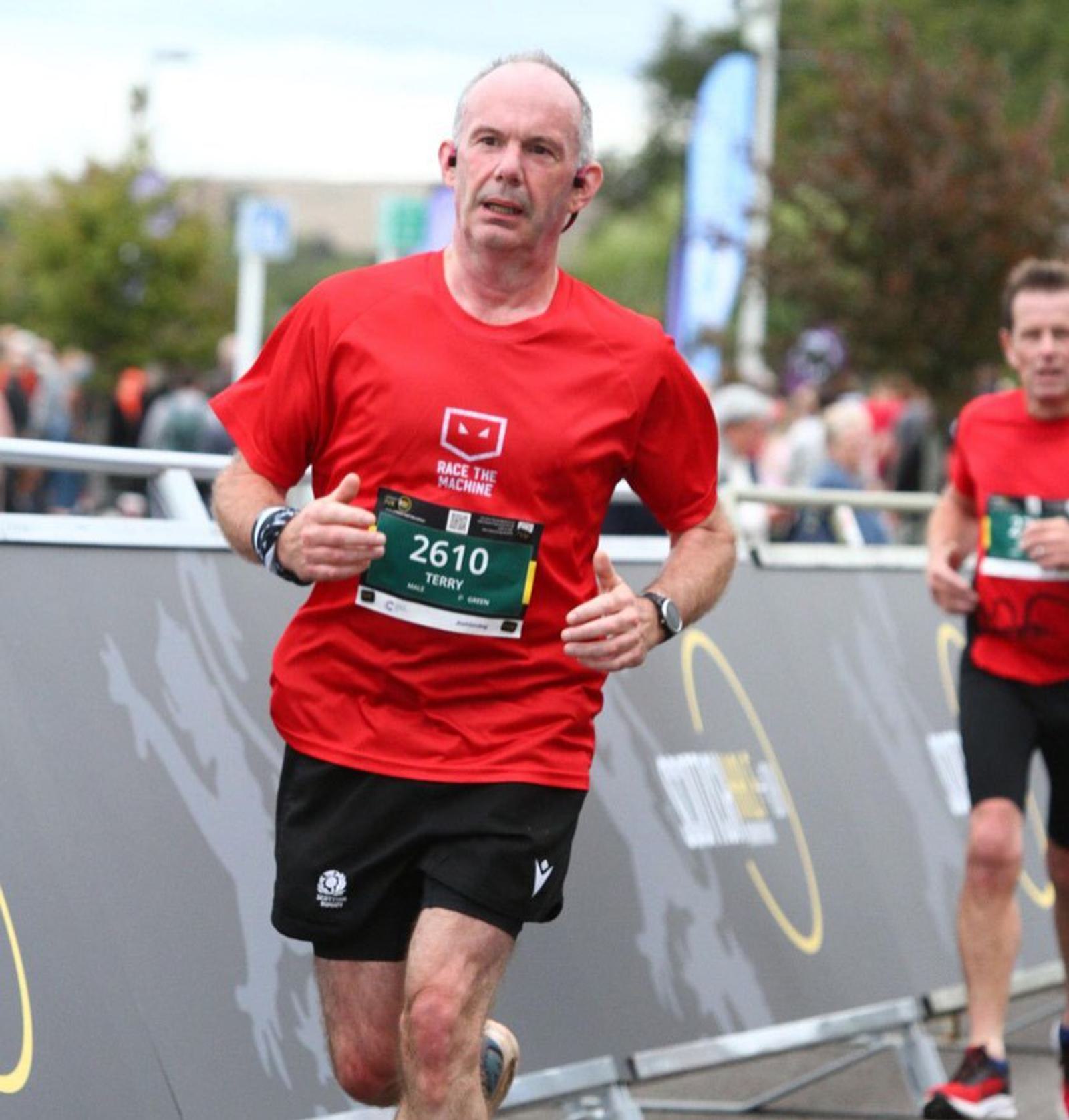 A man in a red shirt and black shorts is running a race, wearing a race bib numbered 2610. He is in an outdoor setting with some greenery and other runners visible in the background. A metal barrier separates the runners from the spectators.