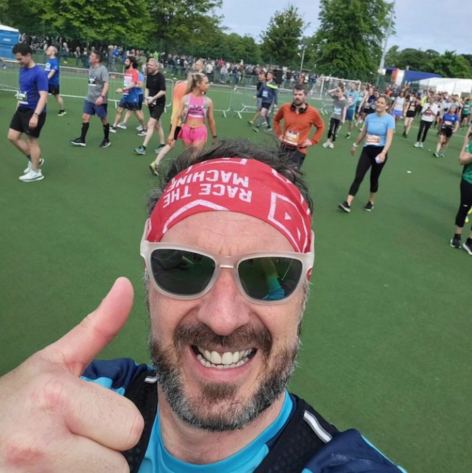 A man wearing a red headband and sunglasses gives a thumbs-up and smiles at the camera. He stands on a green field, surrounded by other people in athletic wear, suggesting they are participating in a race or sporting event. Trees and a filled sky are visible in the background.