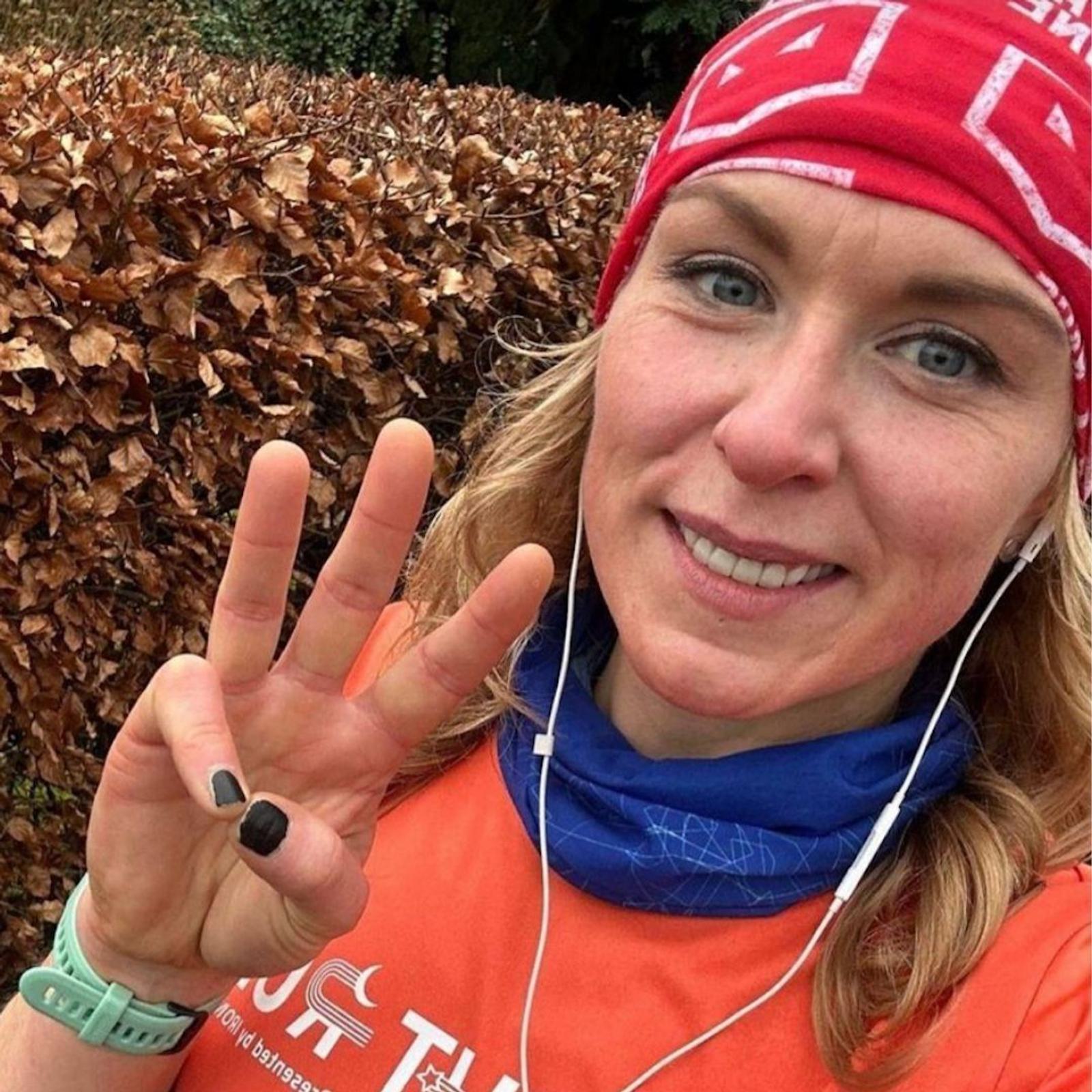 A person wearing a red headband and an orange shirt smiles at the camera. They are holding up three fingers on one hand and have earphones in. There are bushes with dried leaves in the background.