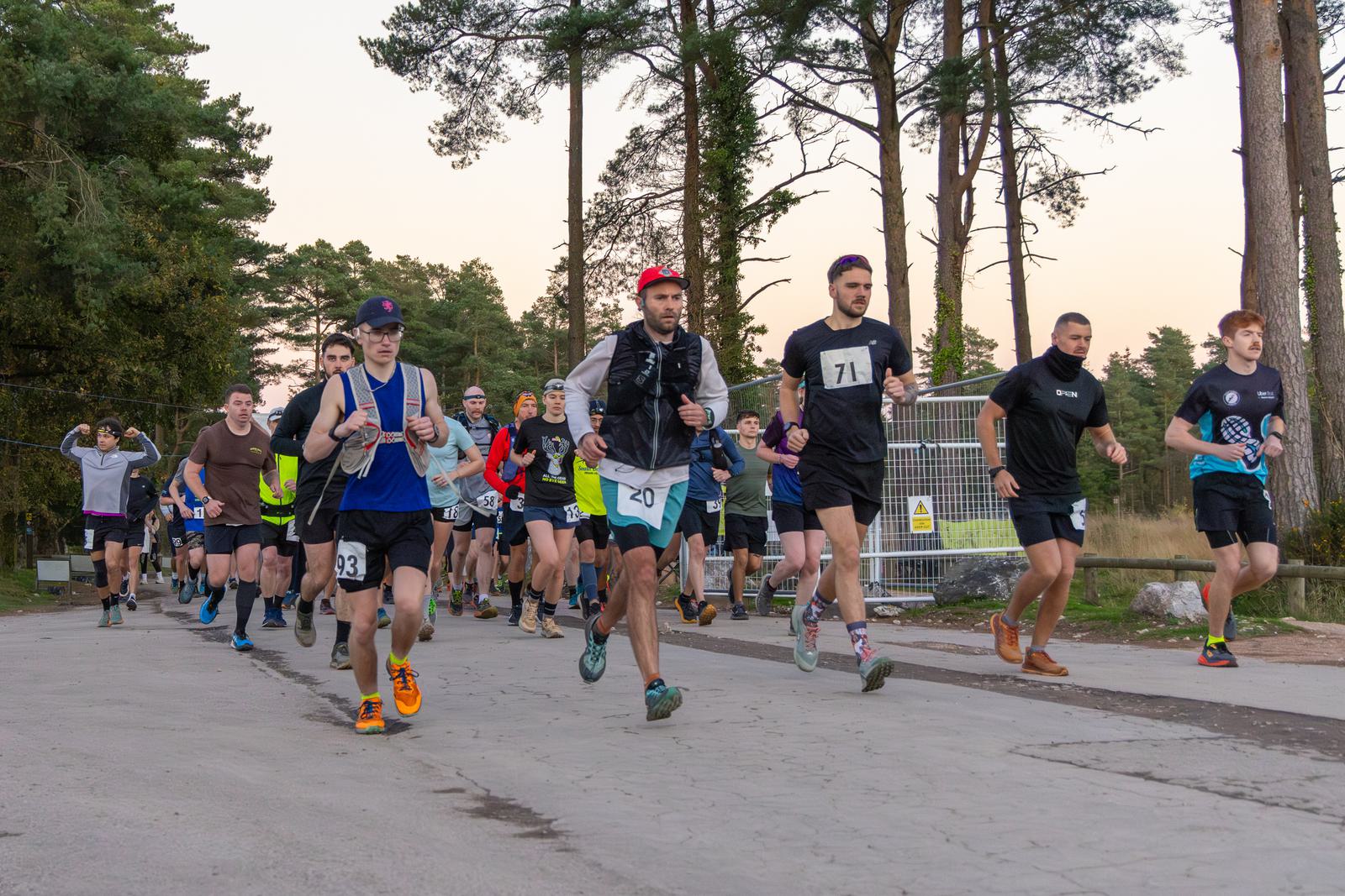 A group of people participate in an outdoor run along a wooded path. Some wear numbered bibs and athletic gear, while others observe. The scene is lively, surrounded by tall trees and a bright sky.
