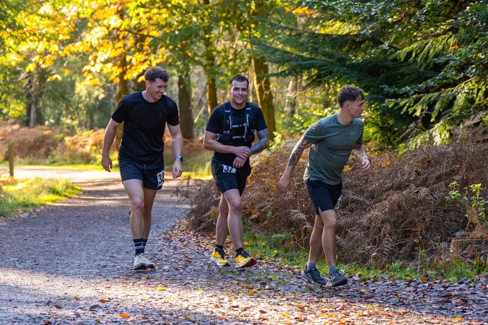 Three men are jogging along a forest trail on a sunny day, surrounded by autumn foliage. They are wearing athletic clothing and appear to be enjoying the outdoor activity.