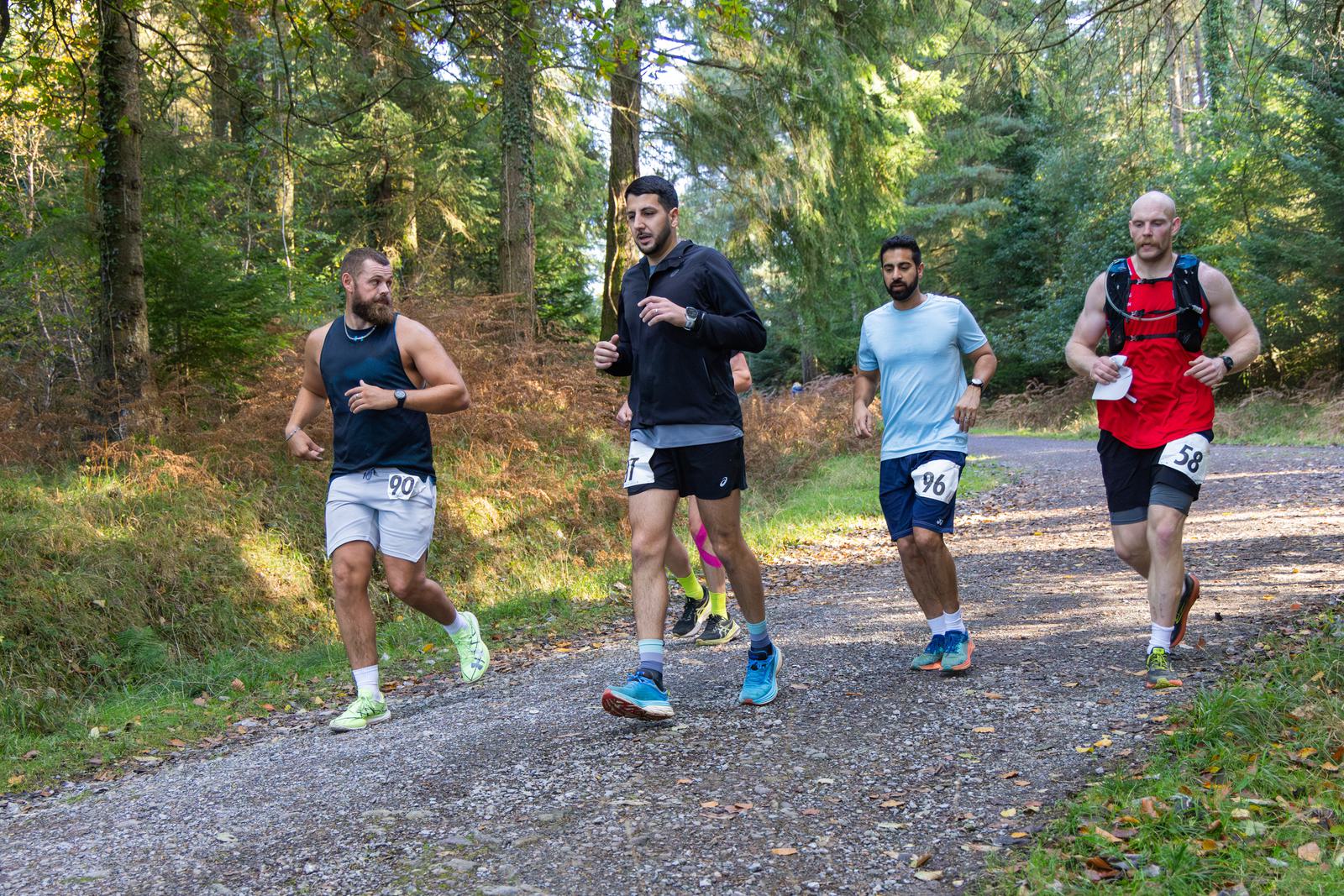 A group of five men jogging on a forest trail. They are wearing running gear with race numbers pinned to their clothes. The path is surrounded by green trees and fallen leaves cover parts of the ground. It is a sunny day.
