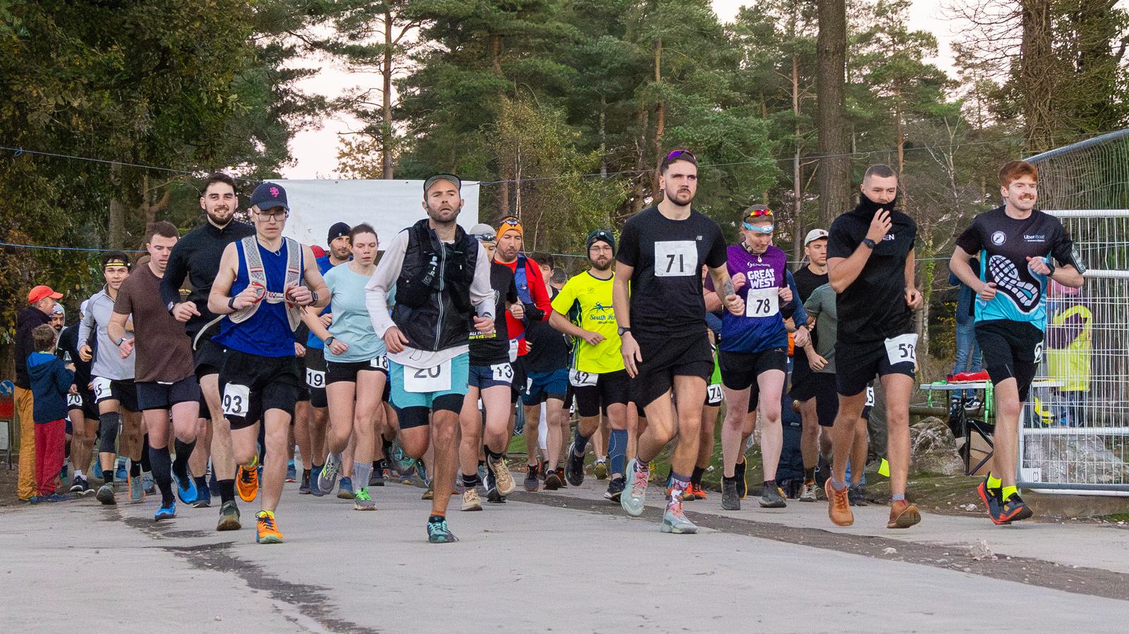 A group of runners in numbered bibs are starting a race on a paved road surrounded by trees. They are dressed in various athletic wear, and the atmosphere is energetic and focused.