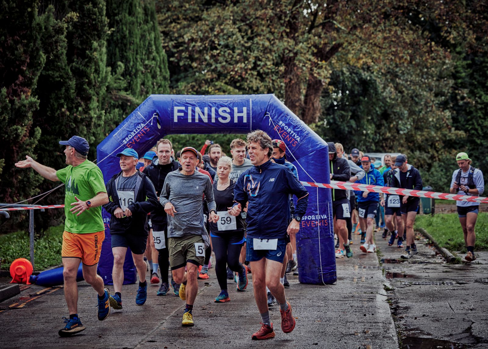 A group of runners, both men and women, cross the finish line of a race event. They appear tired but joyful. The finish line is marked by a blue inflatable arch with "FINISH" written on it. Spectators and other runners are seen in the background under cloudy skies.