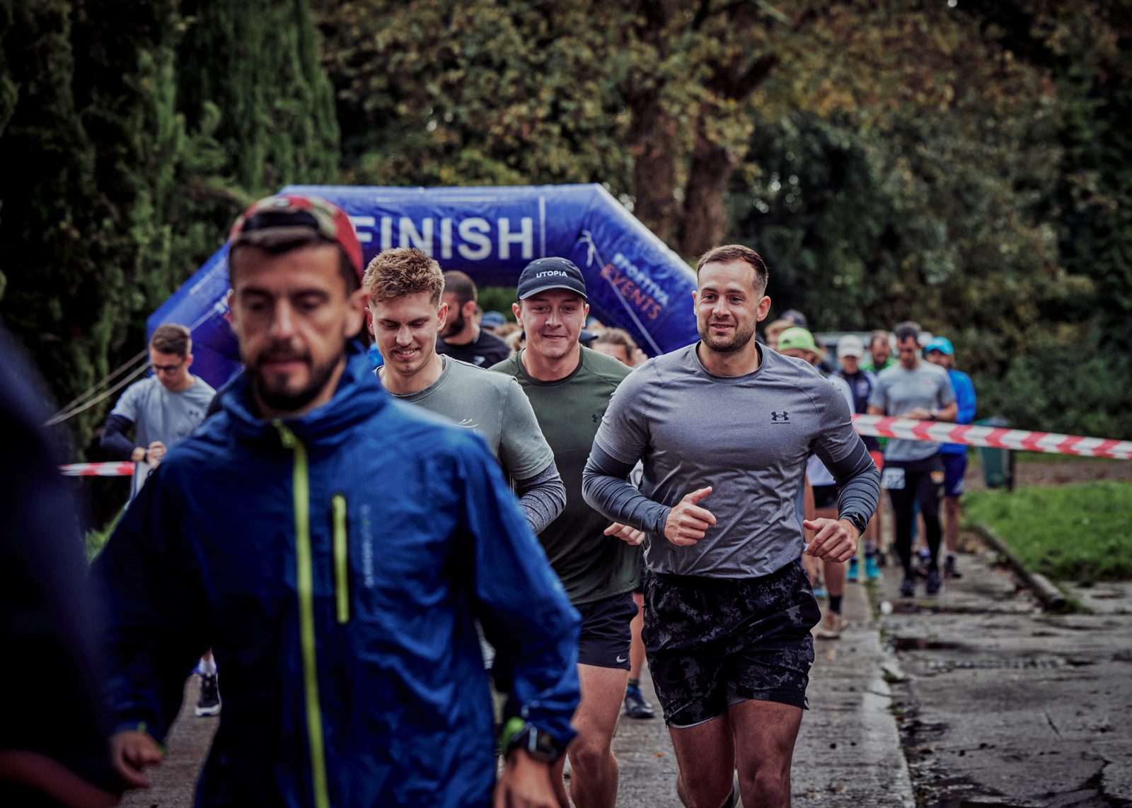 A group of runners approaches the finish line of a race on a cloudy day. Trees and a "Finish" banner are visible in the background. The runners are dressed in athletic gear, with some smiling and others focused. The path is lined with red and white tape.