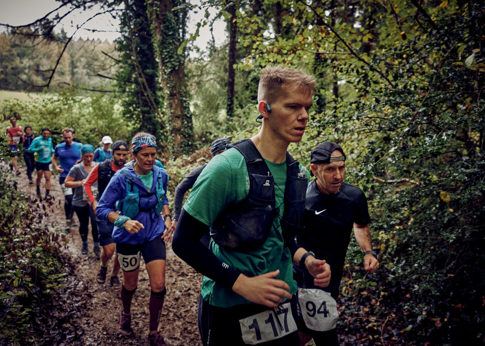 A group of runners competes in an outdoor trail race through a forested area. Participants wear race bibs, hydration packs, and athletic gear suitable for running. The trail is muddy, and the forest is dense with trees and foliage. The atmosphere appears intense.