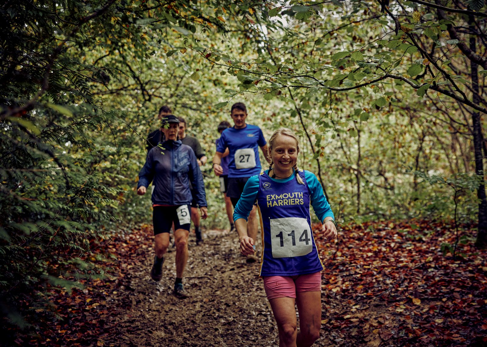 A group of runners, wearing numbered bibs, participates in a trail race through a wooded area. The ground is covered with leaves and the trees are lush with green foliage. The runner in front wears a blue and purple shirt and smiles at the camera.