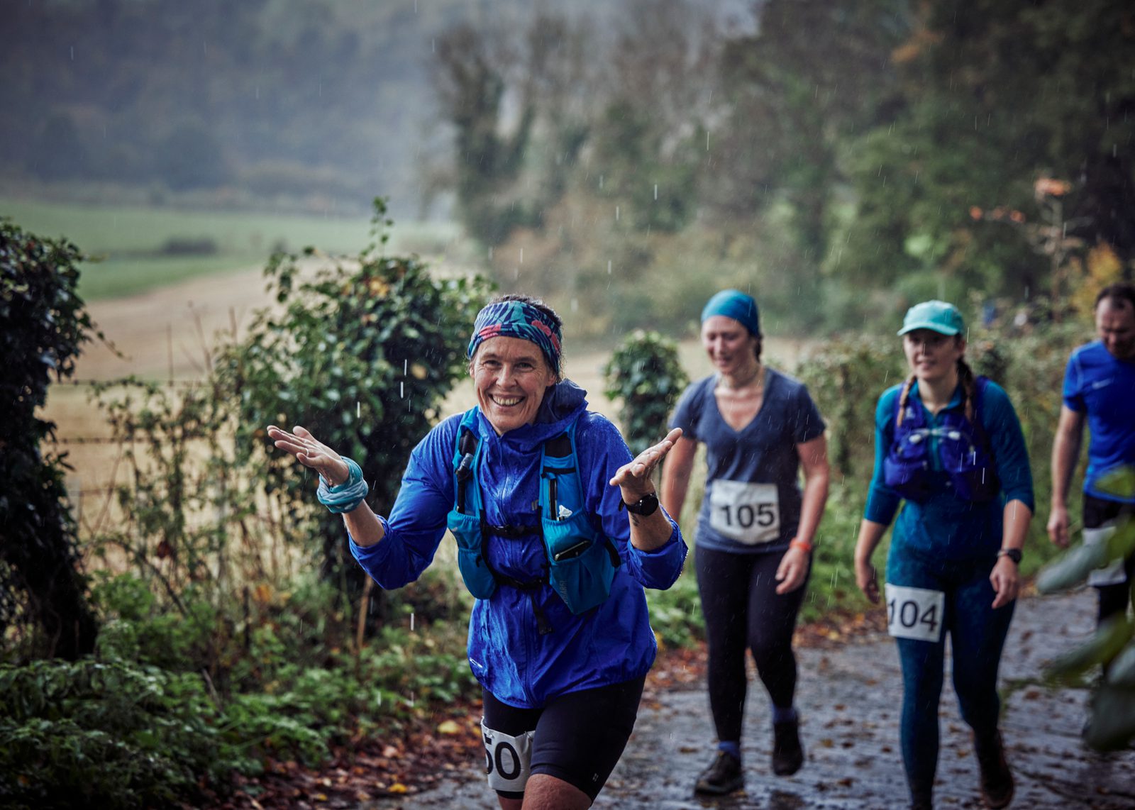 A group of four runners with race bibs on a rain-soaked trail. A smiling woman in the foreground is wearing a blue jacket and headband, raising her hands joyfully. The background features lush greenery and fields, with a misty, overcast atmosphere.