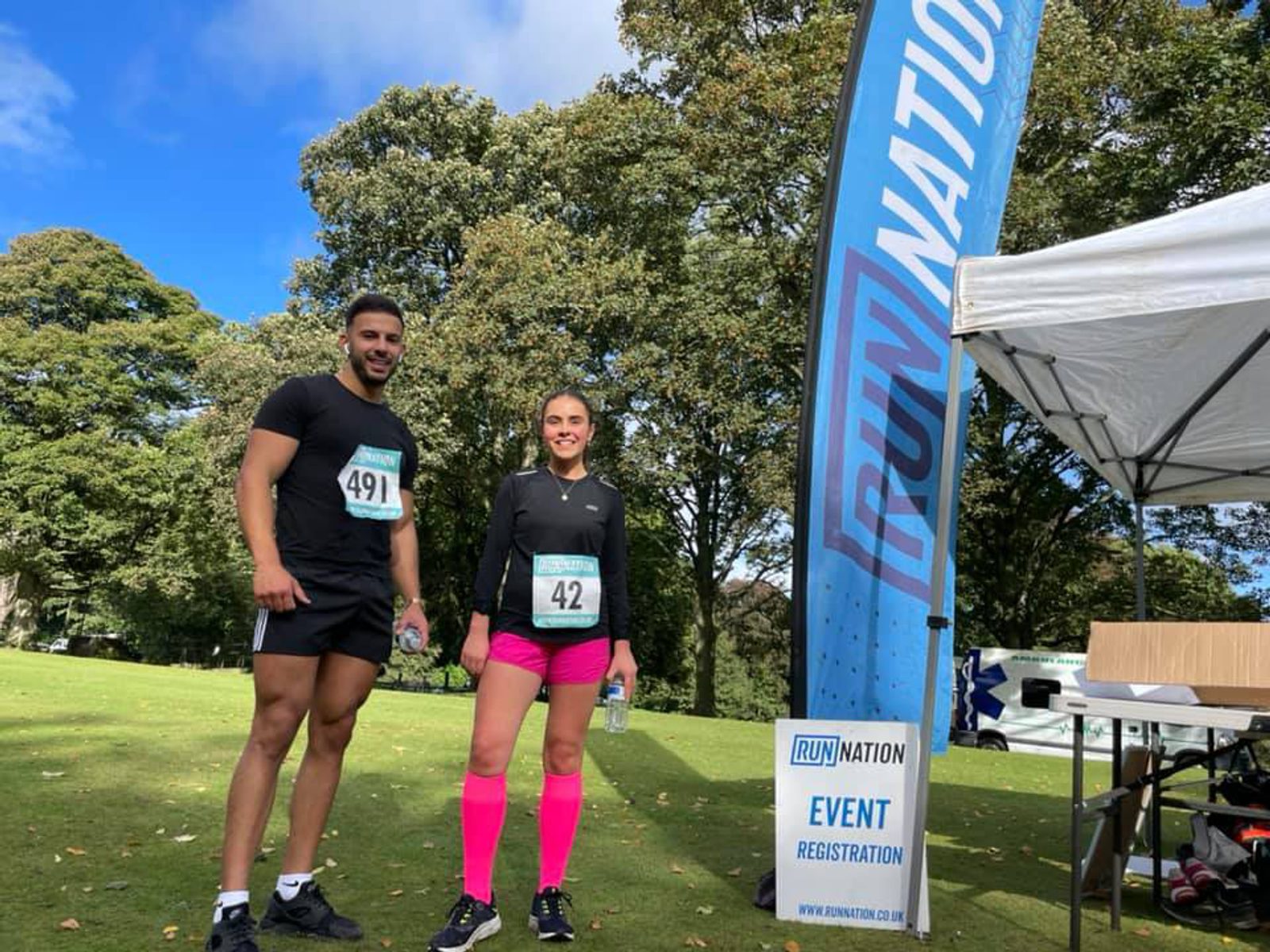 A man and a woman stand in front of a "RUN NATION" sign at an outdoor event. The man is wearing a black shirt and shorts with bib number 491, while the woman is wearing a black shirt, pink knee-high socks, and bib number 42. Trees and a registration tent are visible in the background.