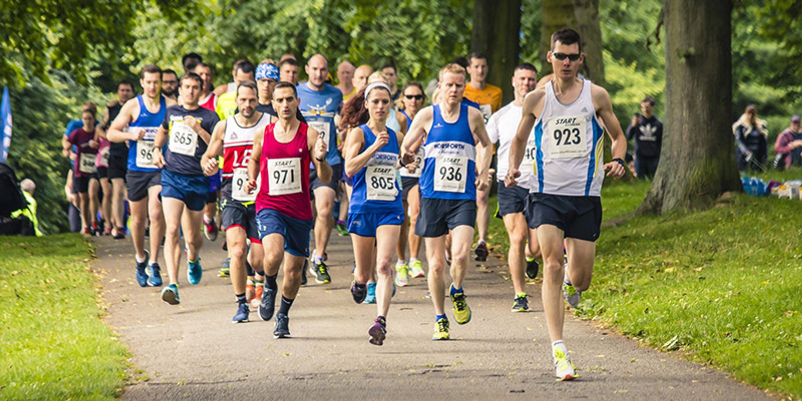 A group of runners is actively participating in a park race event. They are wearing various running gear including numbered bibs pinned to their shirts. The race is taking place on a paved pathway surrounded by lush green trees and grass.