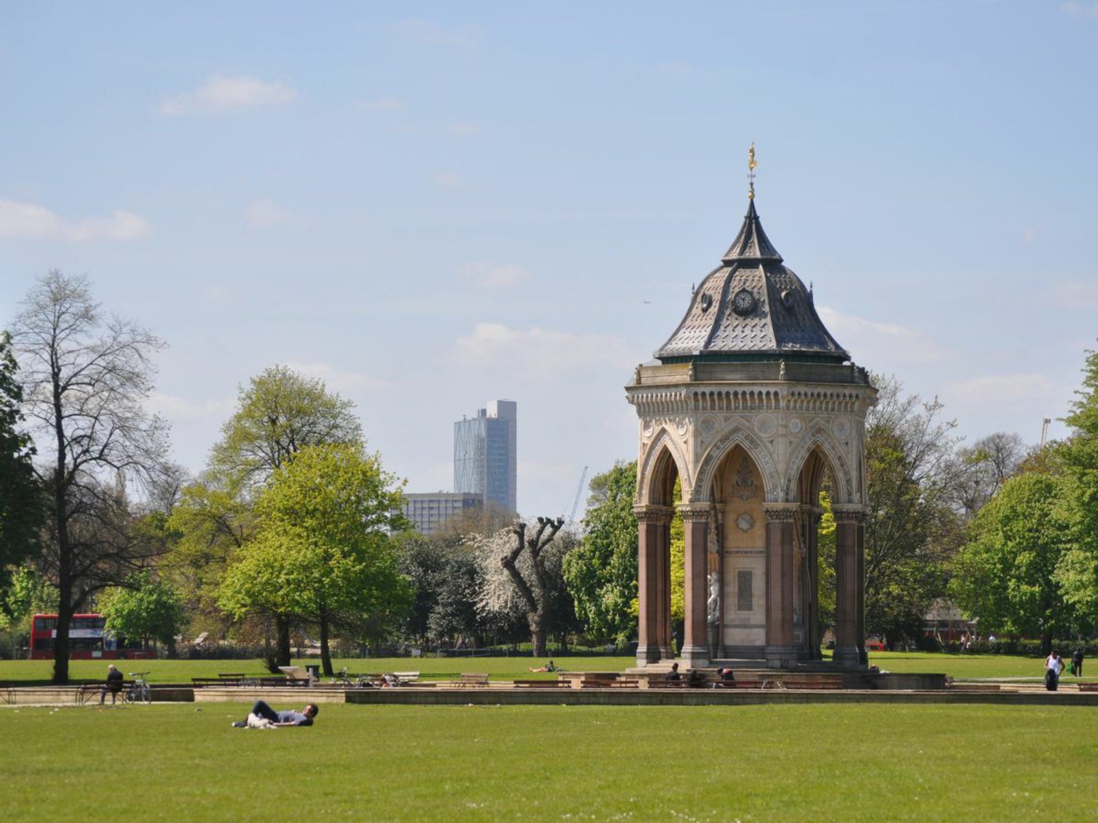 A sunny day in a park featuring a Victorian-style bandstand with intricate architectural details. People are lying on the grass nearby, enjoying the outdoors. Trees and a tall modern building are visible in the background.