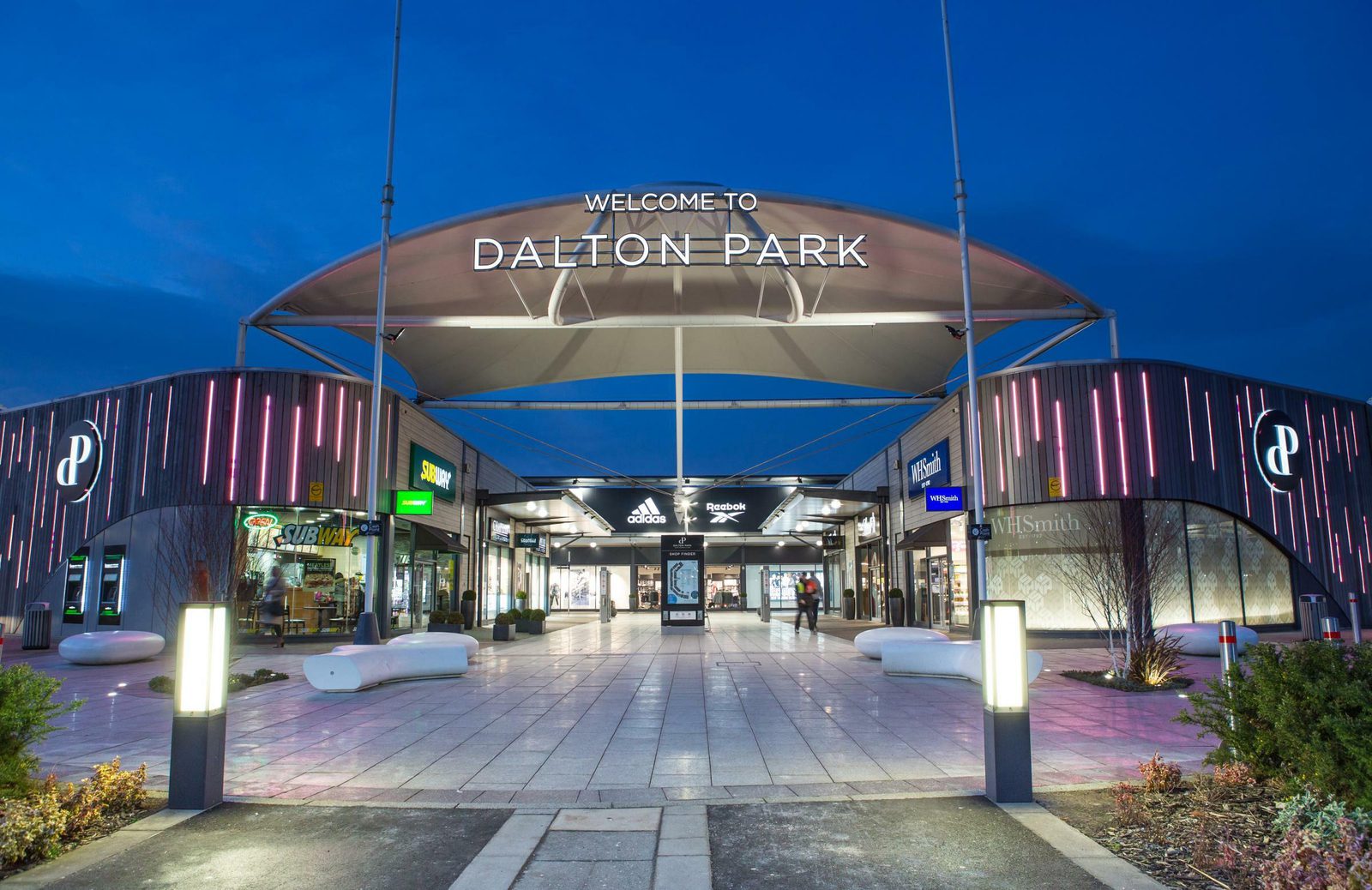 An illuminated, modern shopping center entrance under a twilight sky, featuring the text "Welcome to Dalton Park" on a large canopy structure. Various brand stores, including Subway and Adidas, are visible along with decorative lighting and seating areas.
