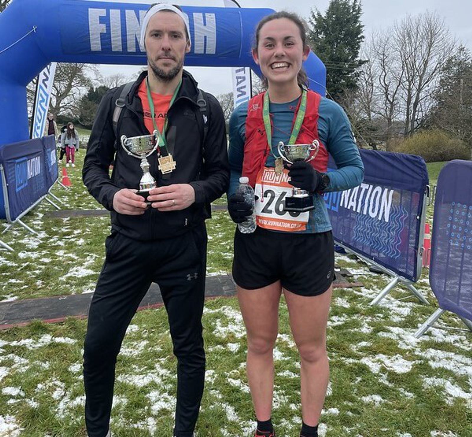 Two runners in winter attire hold trophies and smile at the finish line of a race. The ground is lightly covered in snow, and an inflatable finish arch is visible in the background.
