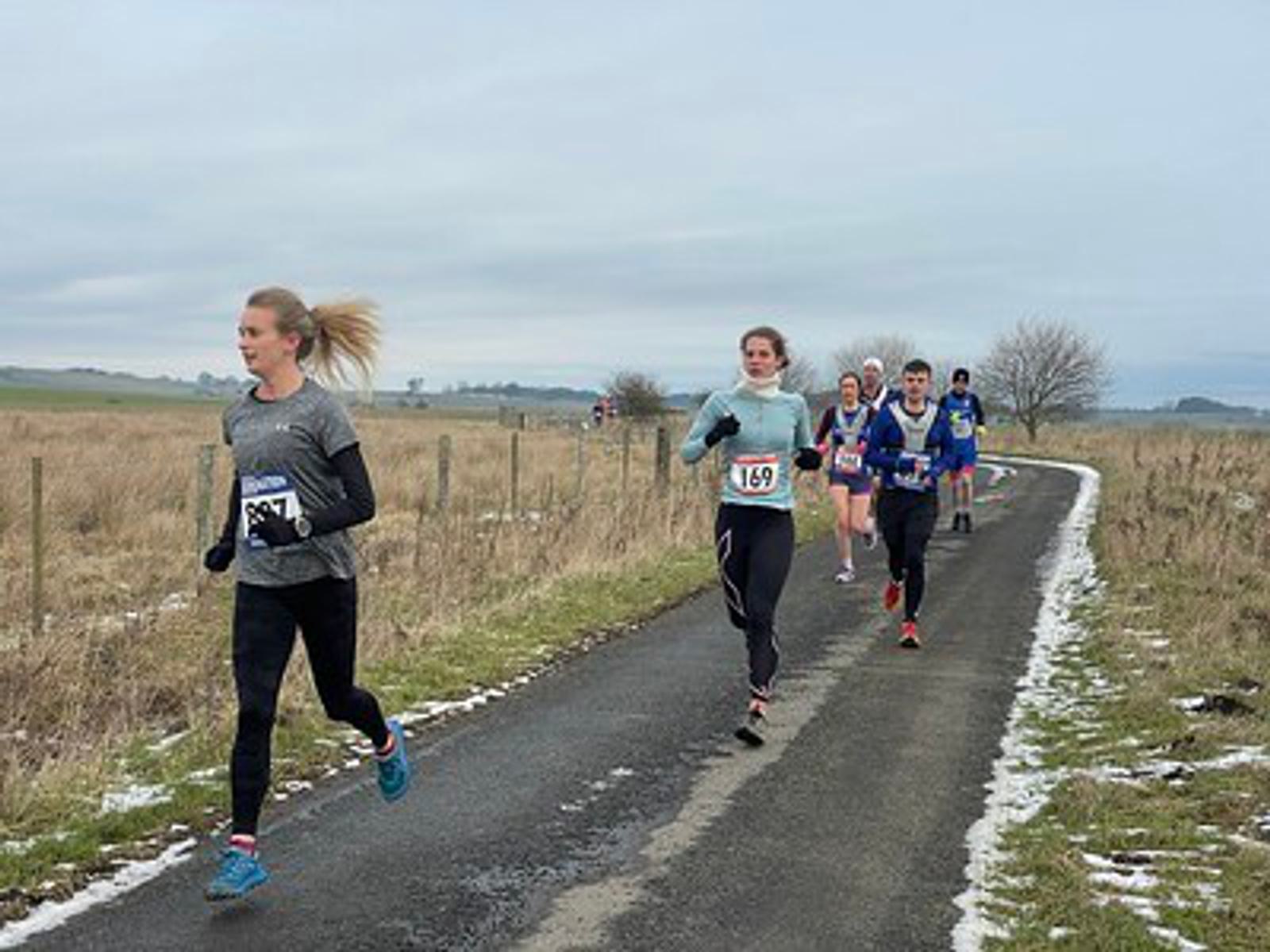 A group of runners competes in a road race on a narrow rural path. The lead runner wears a numbered bib, followed by others, all bundled in winter gear. The surrounding landscape is sparse, with patches of snow and overcast skies.