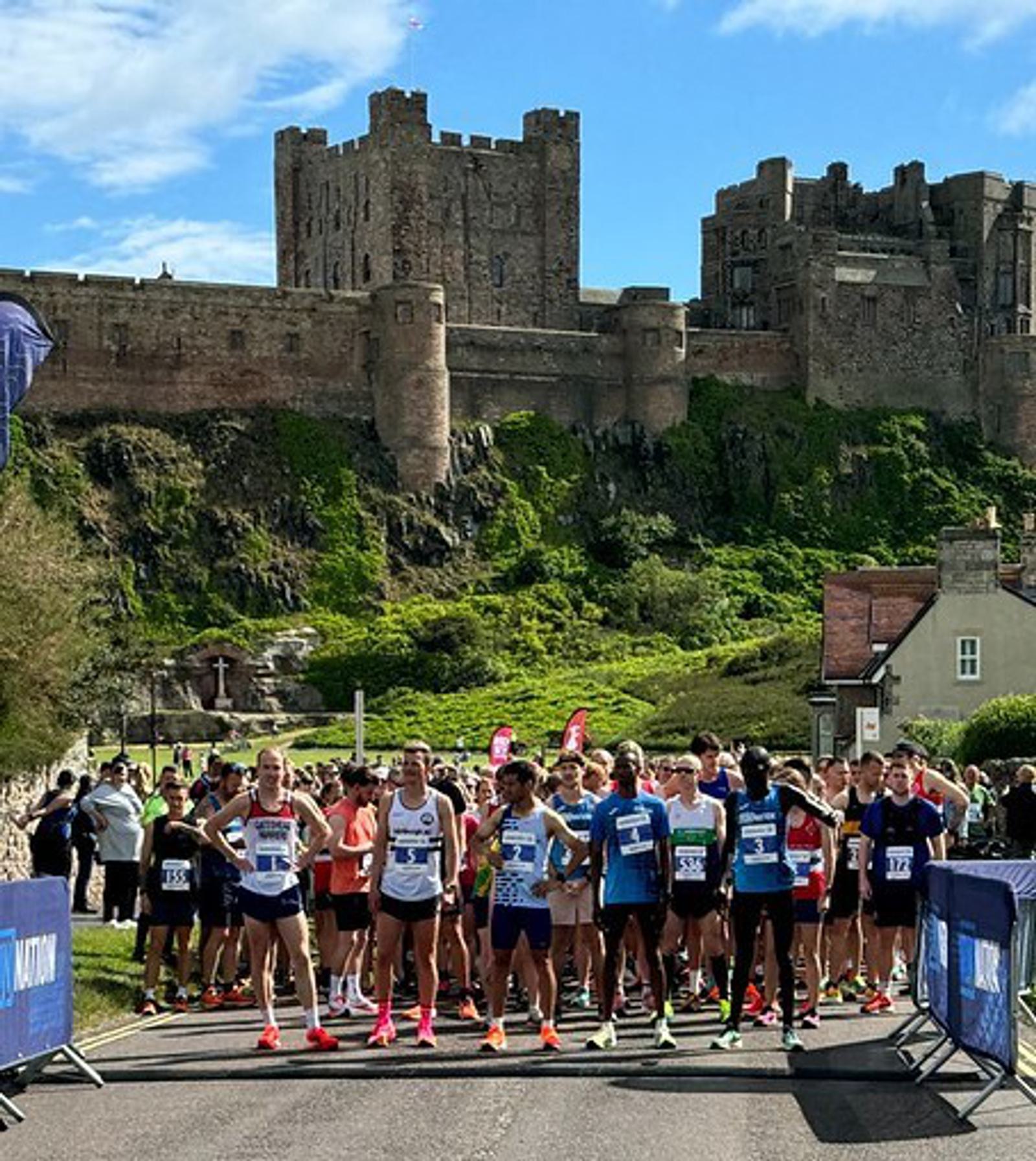 A group of runners stands at the starting line of a race, with an ancient castle on a hill in the background. The day is sunny, and the sky is partly cloudy. Participants wear colorful athletic gear.