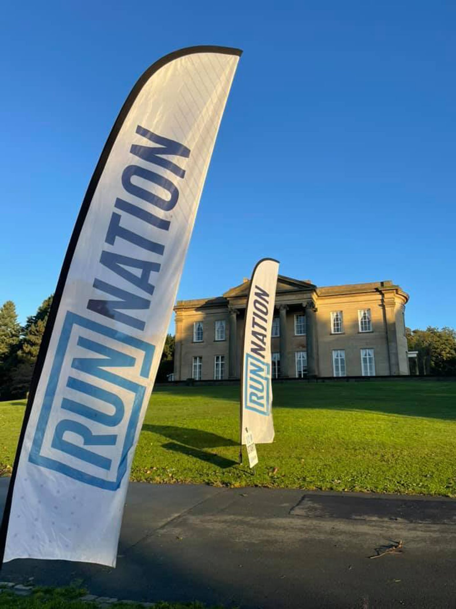 Two vertical flags with the word "Runnation" flutter in the wind on a sunny day. They are positioned on a grassy area in front of an elegant, large building with multiple windows. The clear blue sky and slight shadows suggest it's either morning or late afternoon.
