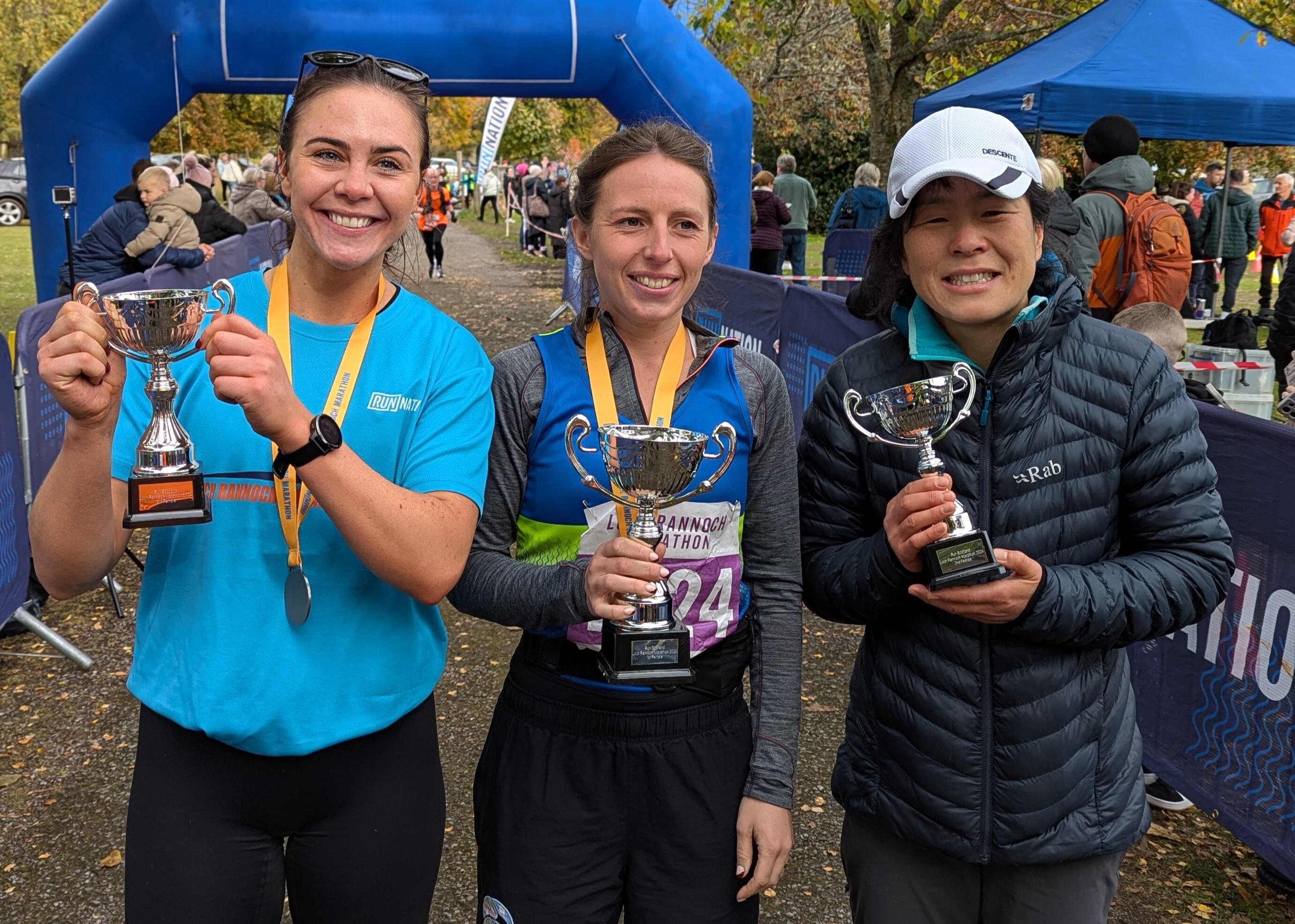 Three women proudly hold trophies at a marathon event. They wear medals around their necks and smile for the camera. An inflated archway and scenic park setting are in the background.