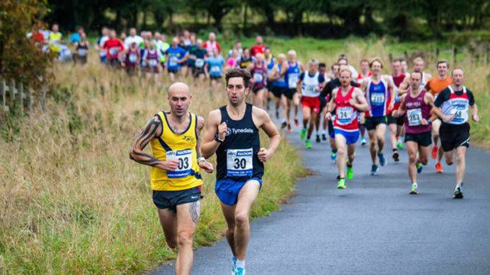 A large group of runners competes in a road race. Two men in the forefront, wearing yellow and black and blue and black outfits, lead the pack, focused and determined. The road is surrounded by greenery, with more runners trailing behind them.