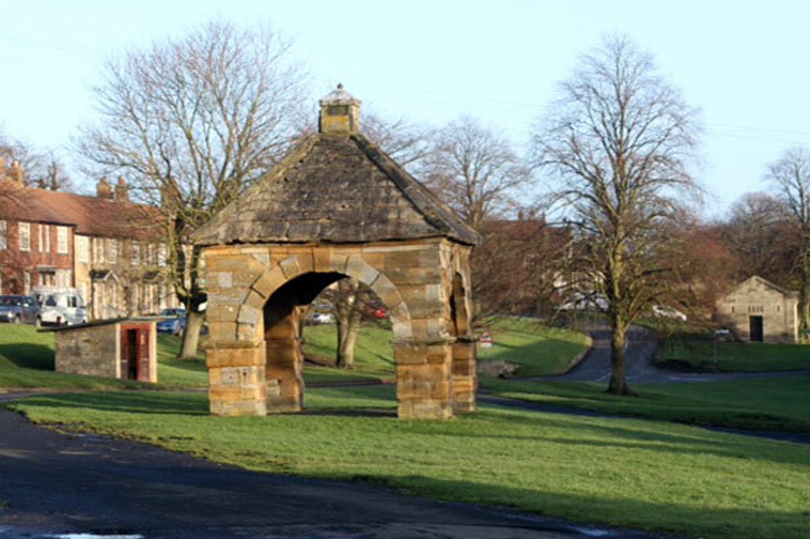 Stone arch structure with a pyramidal roof set in a grassy area surrounded by leafless trees in a small village. In the background, there are houses, a road, and a smaller stone building on a clear day.