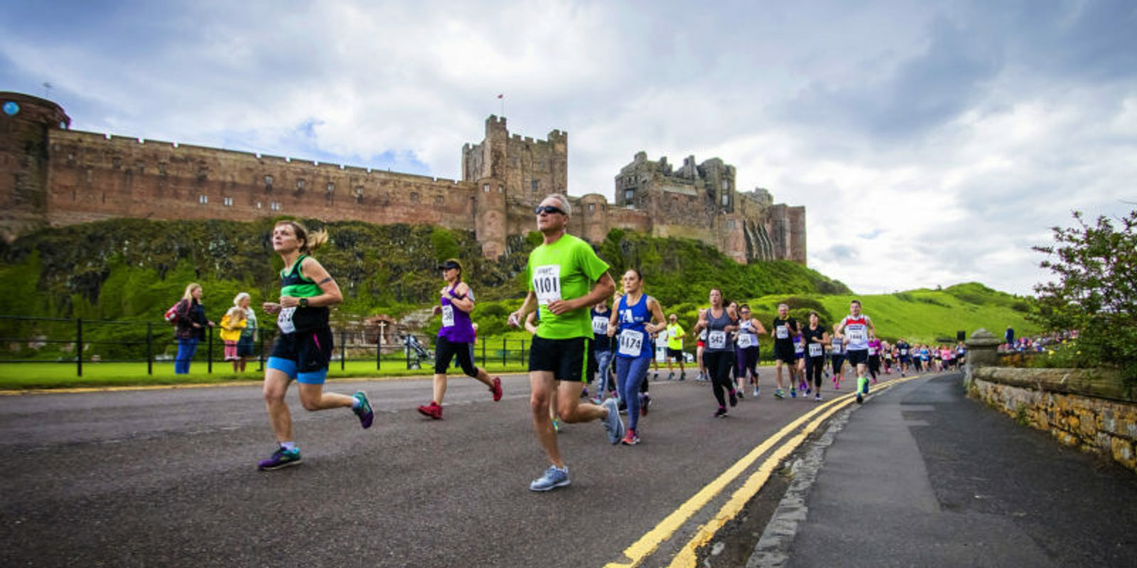 A group of runners participating in a road race near a historic stone castle under a cloudy sky. The runners wear numbered bibs and various athletic attire. Spectators can be seen in the background near the castle grounds.