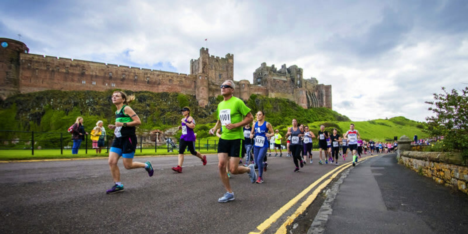 A group of runners participate in a race on a road adjacent to an old, large castle. The sky is cloudy, and some spectators are visible on the side of the road, cheering the runners on. The castle is set on a grassy hill, with its historic walls and towers prominently visible.