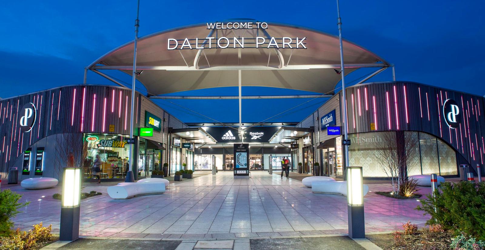 The image shows the entrance to Dalton Park, a shopping mall, under a large, illuminated canopy at twilight. The area is lined with shops, including Subway, Adidas, and Reebok. There is a neon sign and modern white benches along the wide walkway.