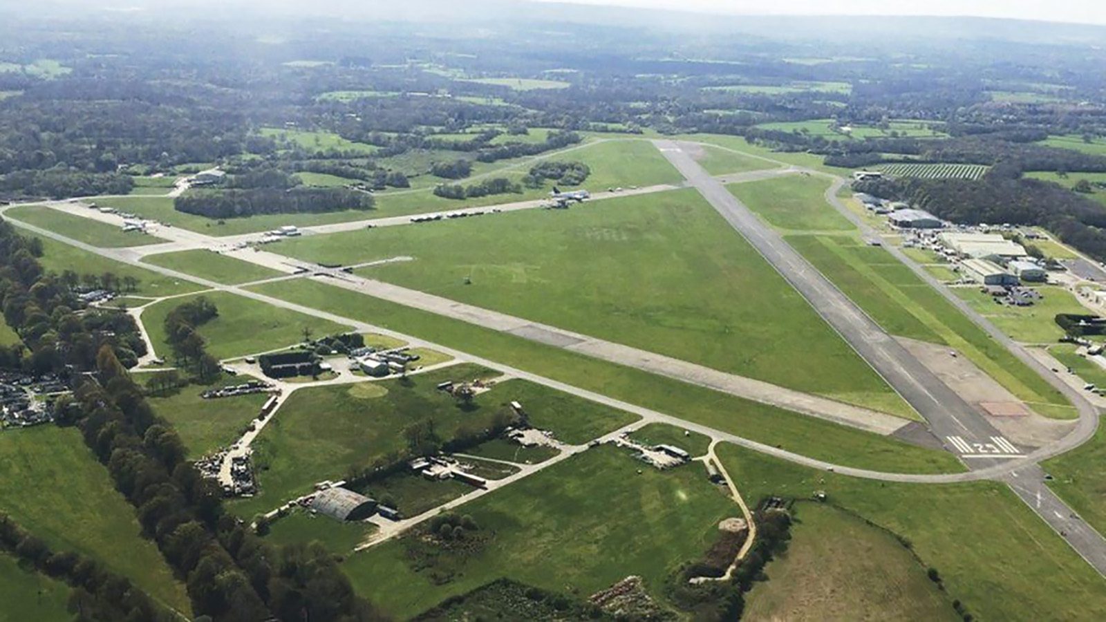 Aerial view of an airfield featuring multiple intersecting runways, taxiways, and adjacent buildings set amidst a vast expanse of green fields and patches of forest. The horizon shows a mix of rural landscapes and distant trees under a clear sky.