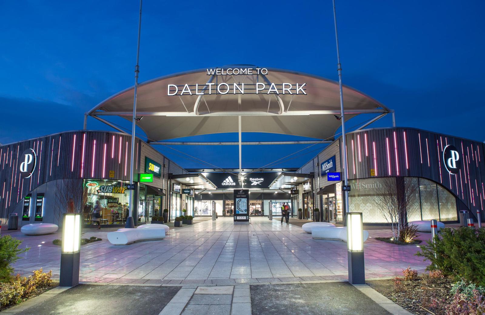 A modern commercial complex with the illuminated sign "Welcome to Dalton Park" at the entrance. The area features various shops, including a Subway and Adidas store, and is adorned with sleek lighting and benches under a canopy roof in an evening setting.