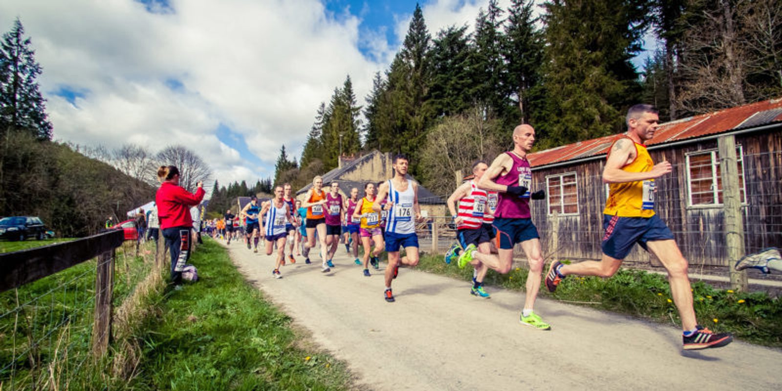 A group of runners competing in a race on a rural road. They are surrounded by trees and rustic buildings. Some participants wear race bibs, sports attire, and running shoes. A few spectators are visible along the side, capturing the event on their phones.