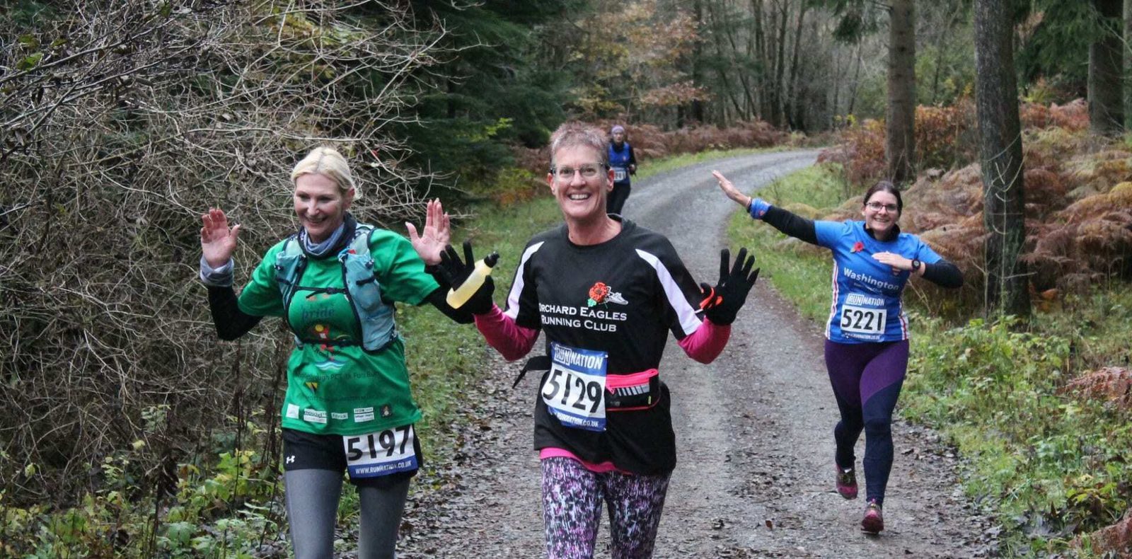 Three runners, all with race numbers, run on a forest trail. Trees and foliage surround them. The runner in the foreground wears a "Road Eagles Running Club" shirt and smiles with raised hands. Two other runners behind also smile and gesture joyfully.