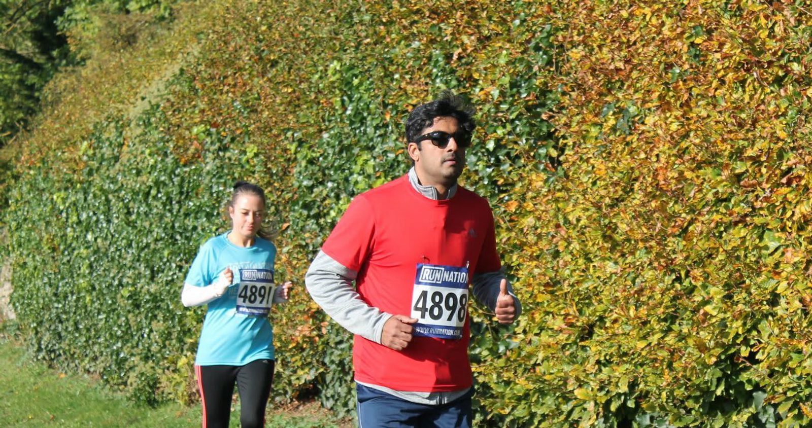 Two people are jogging in a park alongside a tall hedge. The person in front wears a red shirt and dark sunglasses, with a race bib labeled "4898." The runner behind wears a blue shirt and a race bib labeled "4891." Both are focused on the path ahead.