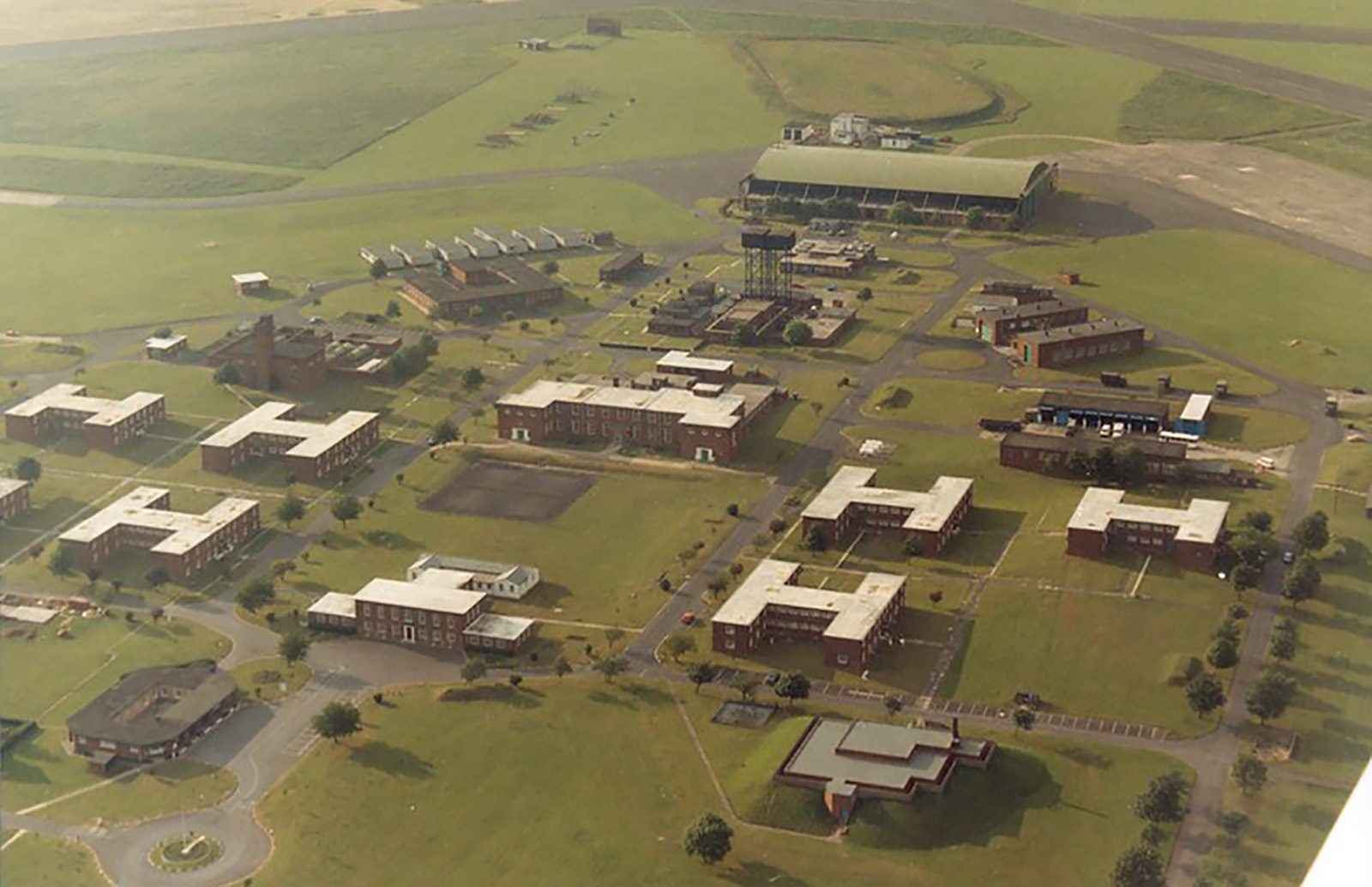Aerial view of a large campus with multiple buildings spread across a manicured lawn. The buildings are connected by roads and pathways, and there are several open grassy areas. In the background, there is a large structure and what appears to be a runway.