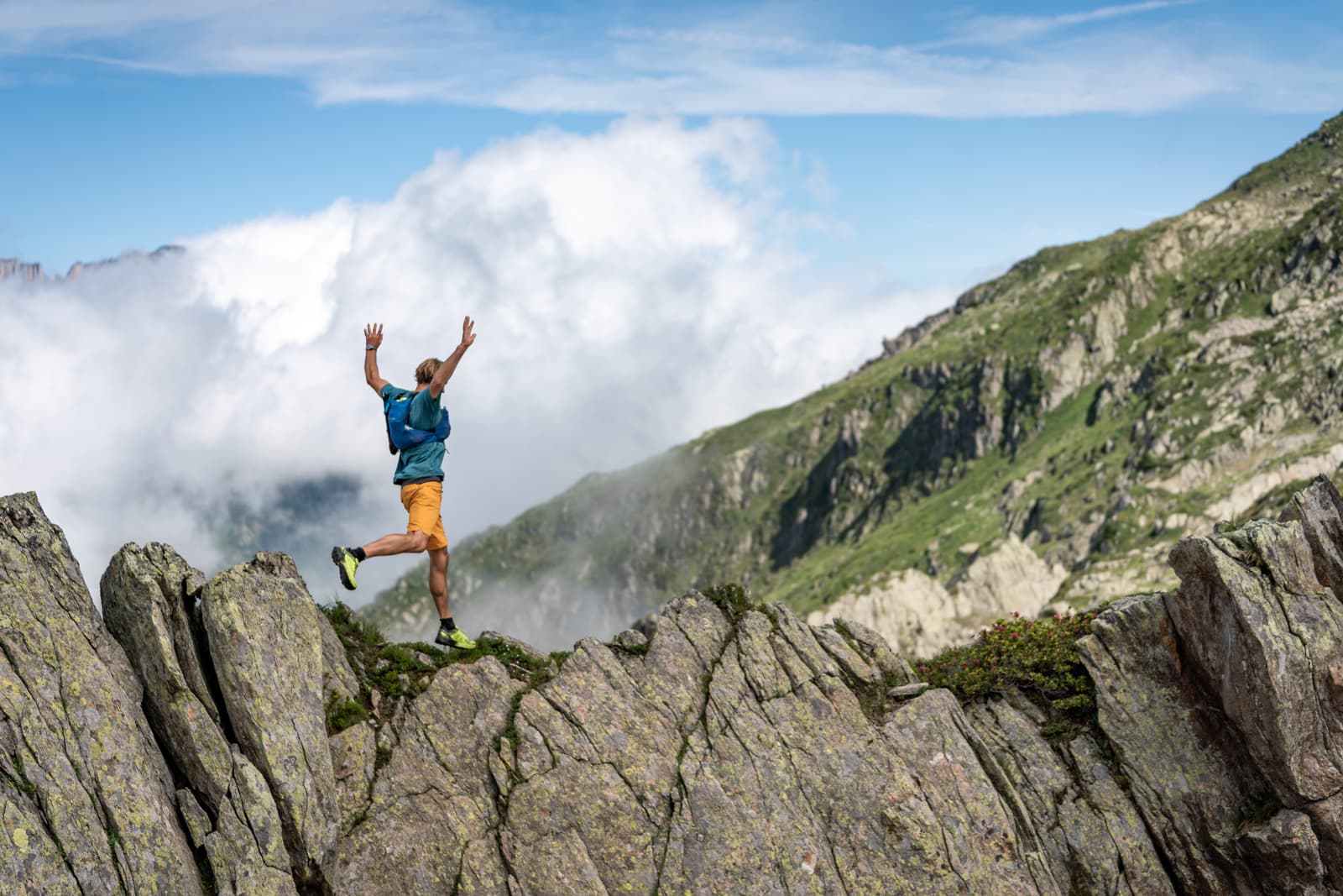 A person in a blue shirt and orange shorts, wearing a backpack, jumps with arms raised over rugged rocks on a mountain ridge. The scene is framed by steep, green slopes and a wide expanse of clouds under a blue sky.