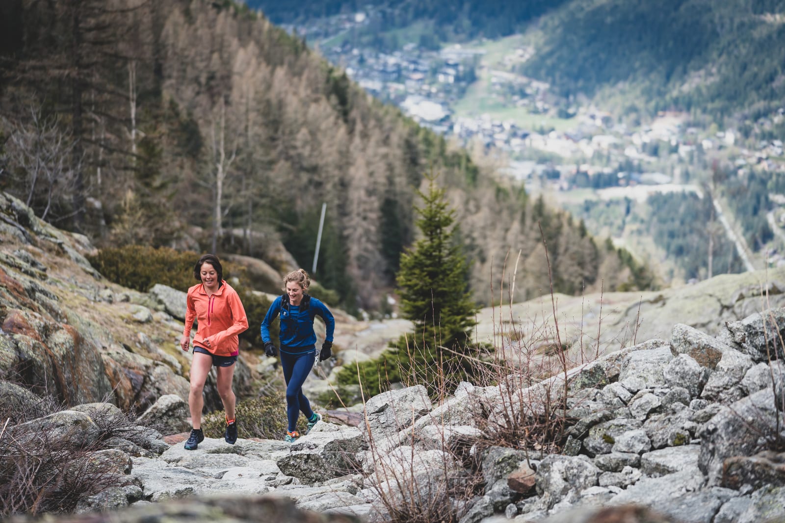Two people are seen hiking on a rocky trail in a mountainous area. One of them is wearing an orange jacket and the other is in blue. The background features a scenic view of a forested mountain slope and a distant village. They appear to be enjoying the outdoor activity.
