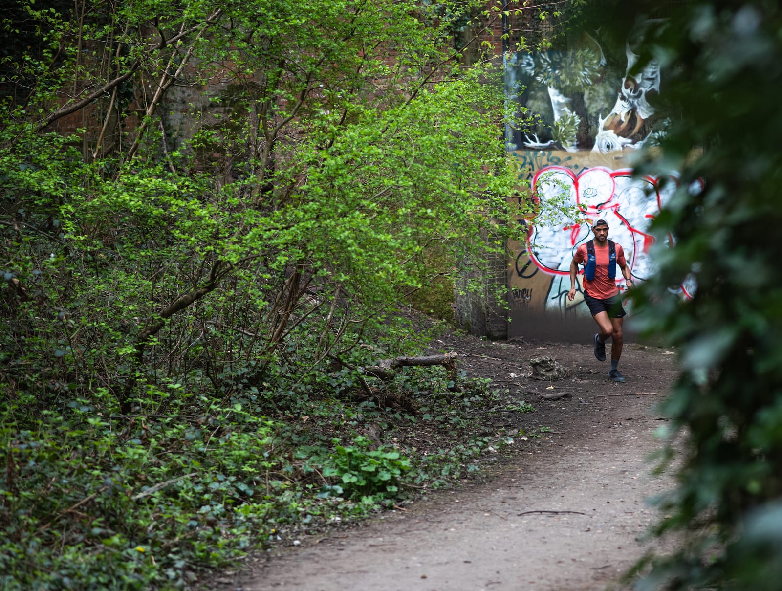 A person jogs down a dirt path through a green forested area, passing a wall covered in colorful graffiti. The greenery surrounding the path is lush with bushes and trees. The person is wearing athletic gear.