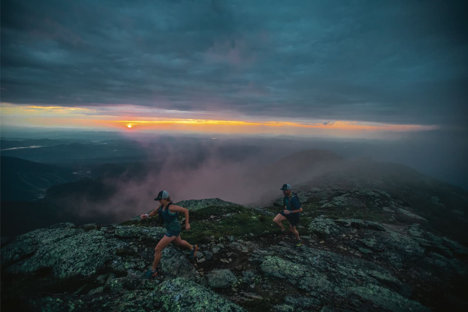 Two people wearing athletic gear ascend a rocky, mountainous trail at dusk. The sky is dark with rolling clouds, and a small glimpse of the setting sun casts a warm, orange hue over the distant landscape, creating a dramatic and moody atmosphere.
