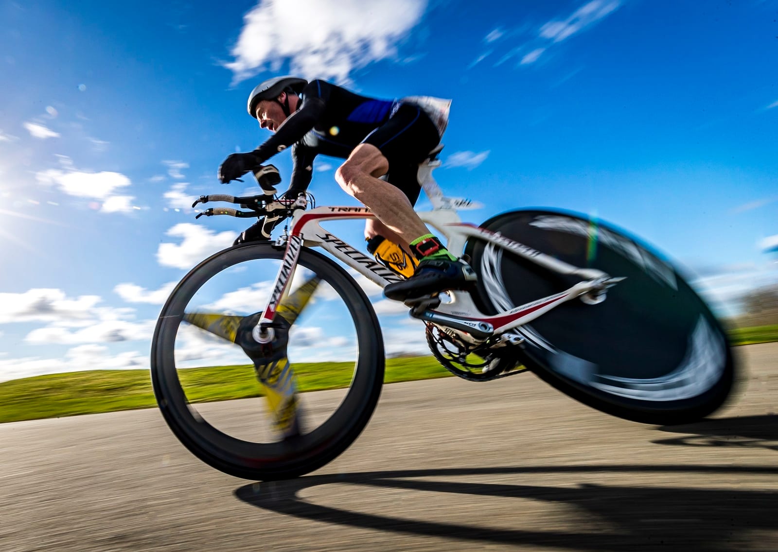 A cyclist wearing a helmet and cycling attire is captured in motion on a road bike. The background is a vibrant blue sky with white clouds, and the ground appears to be a paved surface. The motion blur emphasizes the speed of the cyclist.