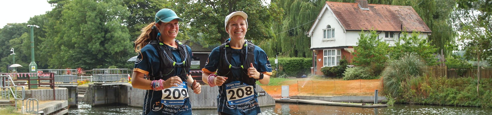 Two people, wearing race bibs 208 and 209, are running along a waterway with a lock and greenery in the background. They appear happy and are dressed in athletic gear with hydration packs. A white house with a red roof is visible on the right side of the image.
