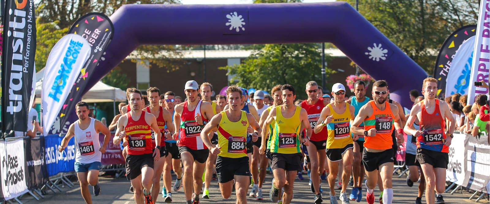 Runners in colorful attire begin a race under a large purple inflatable archway. Spectators line both sides of the street behind barricades, and trees are visible in the background. The atmosphere is energetic under clear skies.