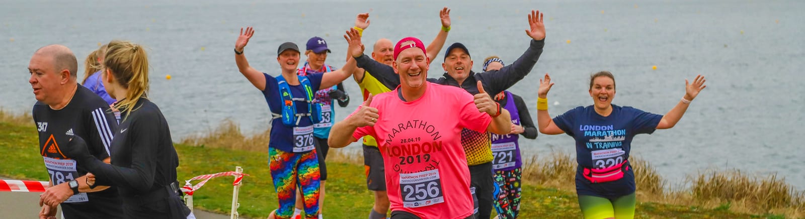 A group of runners in bright clothes are participating in a marathon near a waterfront. They are smiling and waving at the camera, looking joyful. The runner in the center is wearing a pink shirt with "Marathon London" printed on it. The scene is energetic and lively.
