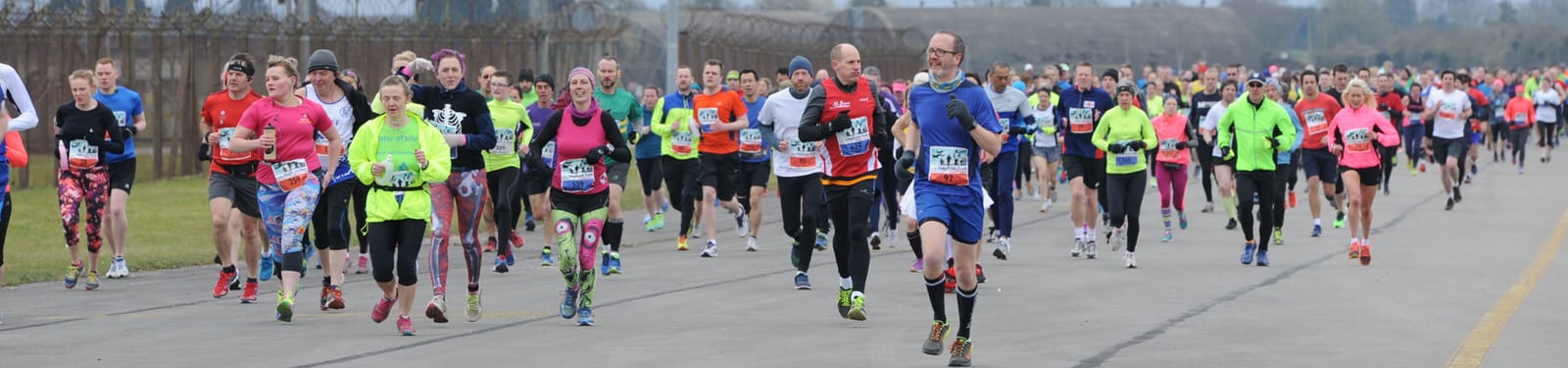 A large group of runners participate in a marathon on a paved road. The runners, both men and women, wear a variety of brightly colored athletic gear. The background is a mix of gray sky and trees, and the atmosphere appears energetic and competitive.