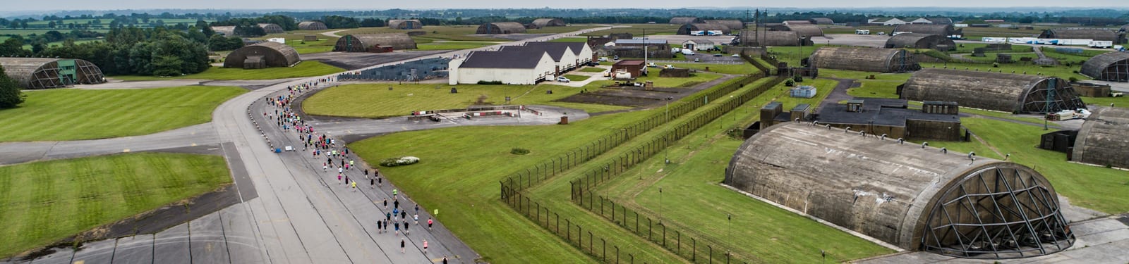 Aerial view of a large, open space with scattered military bunkers and several large warehouse-like buildings. A long road winds through the area, with a group of people walking along it. The landscape is predominantly green, with neatly mowed grass areas.